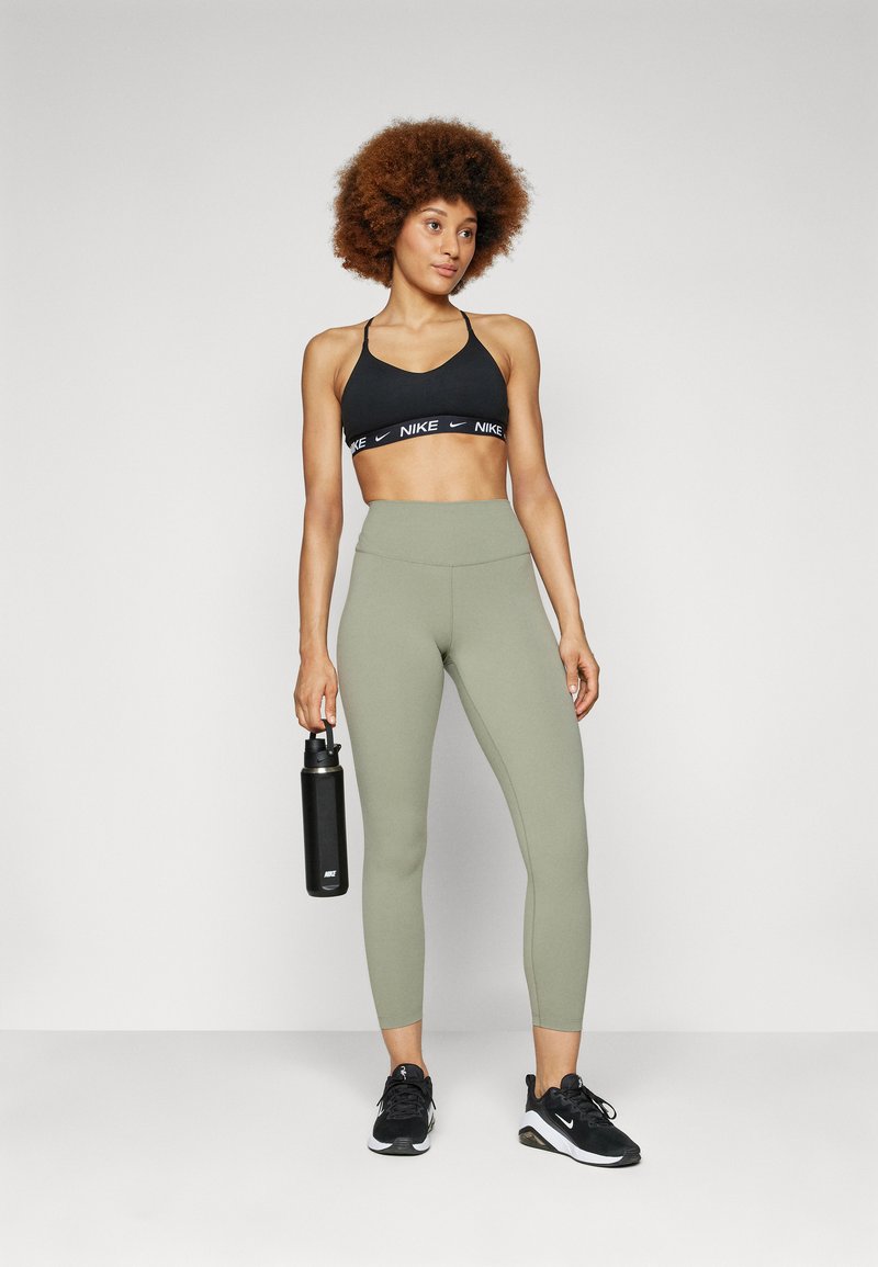 Zwarte sportbeha met logo, olijfgroene hoge leggings, zwarte sportschoenen en een zwarte waterfles in de hand.