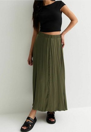 Frau trägt ein schwarzes kurzes Oberteil, einen olivgrünen plissierten Maxirock und schwarze Sandalen mit offenen Zehen, stehend vor einem schlichten weißen Hintergrund.