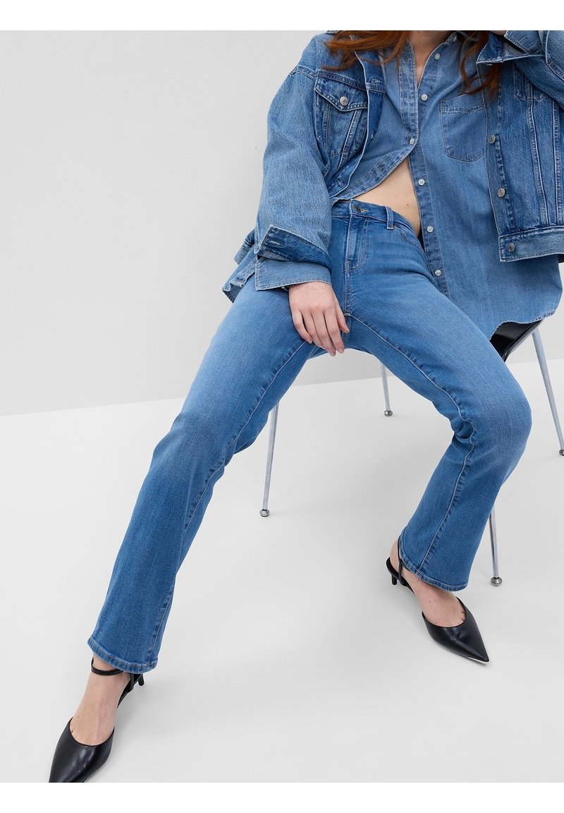 Femme assise sur une chaise portant une veste en jean bleue, un jean bleu et des escarpins noirs à bout pointu avec brides à la cheville.