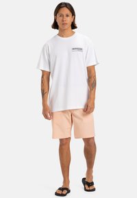 Camiseta blanca de manga corta con un logo en el pecho, combinada con pantalones cortos rosa claro y sandalias negras, creando un atuendo casual.