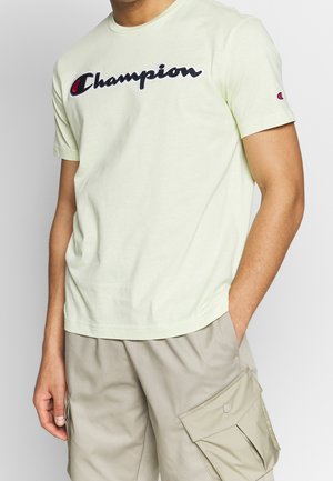 Homme portant un t-shirt Champion jaune clair avec un logo noir et un short cargo beige, debout avec une main dans la poche.