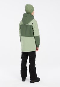 Giacca verde con cappuccio e striscia orizzontale verde scuro, polsini elasticizzati, abbinata a pantaloni neri e stivali neri, vista da dietro.