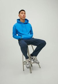 Blauwe hoodie met trekkoorden, marineblauwe broek en grijze sneakers met witte veters, zittend op een kruk tegen een neutrale achtergrond.