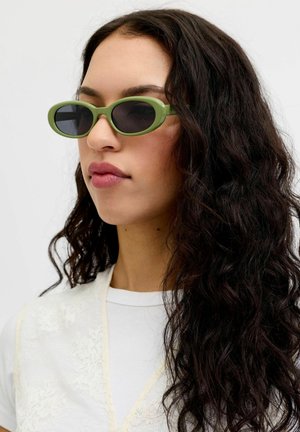Kvinde med langt krøllet mørkt hår iført ovale grønne solbriller og en hvid top mod en ensfarvet lys baggrund.
