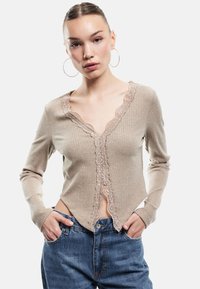 Beige geripptes Strick-Cardigan mit V-Ausschnitt, Spitzenbesatz entlang der Vorderseite und langen Ärmeln. Getragen mit hoch taillierten blauen Jeans.