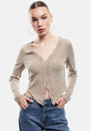 Beige geripptes Strick-Cardigan mit V-Ausschnitt, Spitzenbesatz entlang der Vorderseite und langen Ärmeln. Getragen mit hoch taillierten blauen Jeans.