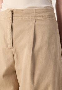 Beige katoenen broeken met plooien, een soepele textuur en subtiele stikseldetails bij de tailleband en zakken.