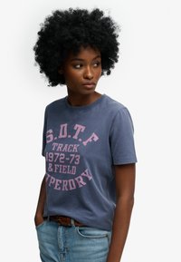 Blauw katoenen T-shirt met roze grafische tekst, ronde hals, korte mouwen en een losse pasvorm. Draagt lichtblauwe spijkerbroek en een bruine riem.