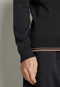 Czarny dziany sweter z prążkowanym mankietem i krawędzią z cienkimi, poziomymi paskami w kolorze brązowym i beżowym. Materiał wydaje się gładki i miękki.