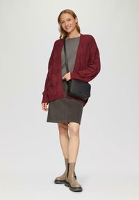 Burgunderfarbener Strickcardigan mit Zopfmuster, graues Oberteil, dunkelgrauer Cordrock und beige Ankle Boots. Schwarze Umhängetasche mit strukturiertem Finish.