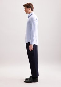 Camicia azzurra chiara a maniche lunghe con polsini abbottonati, fessure laterali, abbinata a pantaloni scuri e scarpe nere, vista di lato.