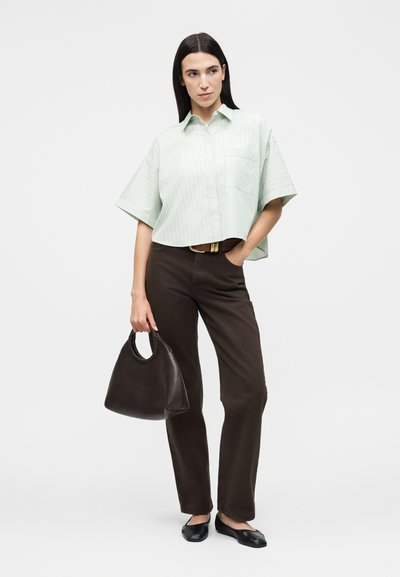 Γυναίκα με ανοιχτοπράσινο ριγέ κοντό πουκάμισο, σκούρο καφέ παντελόνι, μαύρες μπαλαρίνες, κρατώντας μεγάλη μαύρη τσάντα, στέκεται μπροστά σε ένα απλό φόντο.