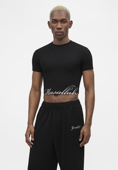 Άνδρας με κοντά ξανθά μαλλιά που φοράει εφαρμοστή μαύρη κοντή μπλούζα με την επιγραφή "Masallah" και φαρδιά μαύρη παντελόνια με λευκό κείμενο στο αριστερό μηρό.