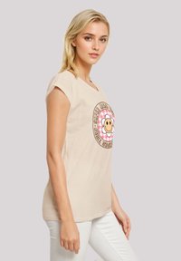 Beige short-sleeve t-shirt featuring a colorful circular graphic with a smiley face and flowers, paired with white jeans.