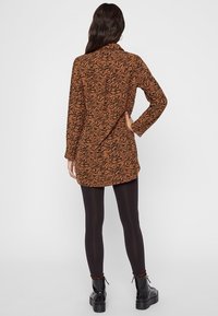 Maglietta lunga a strisce arancioni e nere con colletto abbottonato, indossata sopra leggings neri e stivali neri chunky. Tessuto texture, vestibilità comoda.