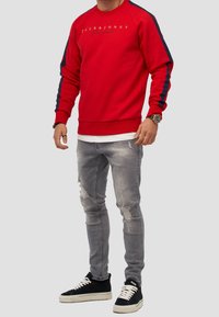 Felpa rossa con strisce laterali blu navy, dotata di un logo sul petto. Indossata con jeans chiari strappati e sneakers nere. Texture liscia.