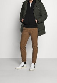 Giubbotto piumino verde oliva, felpa nera con zip, pantaloni marroni aderenti e sneakers bianche con dettagli neri. Texture liscia, vestibilità casual.
