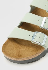 Svijetlozeleni sandal s tri podesiva remena, metalnim kopčama i plutenom iznutricom. Ima teksturiranu smeđu unutrašnjost i crnu vanjsku stranu.