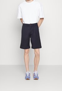 Person som bär en vit T-shirt, mörka knälånga shorts och blåa och rosa sneakers står mot en enfärgad ljus bakgrund.