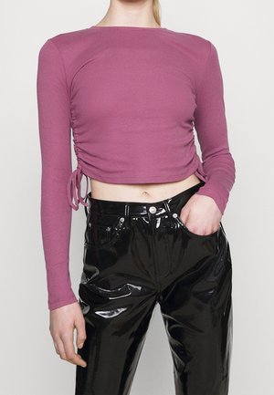 Frau trägt ein mauvefarbenes langärmeliges Crop-Top mit seitlichen Bändern und schwarze glänzende Vinylhosen, eine Hand in der Tasche.