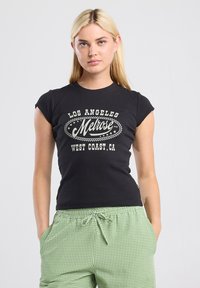 Black ribbed t-shirt featuring a central white graphic text print. Short sleeves and a fitted shape. Green checkered pants visible below.