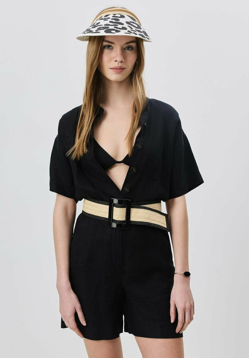 Schwarzer Leinen-Jumpsuit mit kurzen Ärmeln und tiefem Ausschnitt, tailliert mit einem breiten cremefarbenen Gürtel und einer schwarzen Schnalle. Mit einem gemusterten Visierhut.