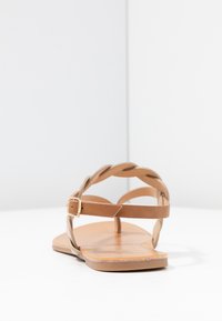Brune lædersandal med et flettet stropdesign, flad sål og justerbar ankelspænde. Har en glat overflade og minimalistisk stil.