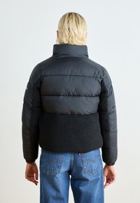 Svart boblejakke med en teksturert svart fleecepanel i midjen, høy krage og elastiske mansjetter, brukt med blå denimjeans.