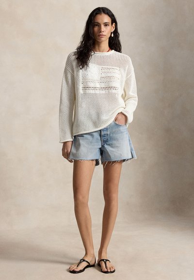 Γυναίκα με λευκό πλεκτό πουλόβερ, πολύχρωμα ανοιχτό μπλε τζιν σόρτς, μαύρες σανδάλες, στεκόμενη μπροστά σε μια απλή μπεζ φόντο.