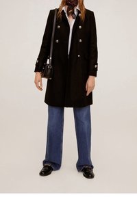 Abrigo negro de doble botonadura con botones plateados, llevado sobre una camisa blanca y jeans de pierna ancha azules, complementado con una bufanda estampada y zapatos negros.