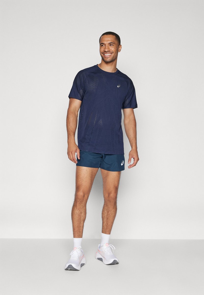Marinefarbene Sport-T-Shirt mit kurzen Ärmeln, Netzstruktur und Logo. Kombiniert mit türkisfarbenen Shorts und weißen Sportschuhen mit rosa Akzenten.