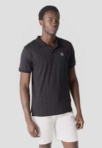Jeune homme aux cheveux courts et bouclés portant un polo noir à pois et un short blanc, debout devant un fond gris uni.