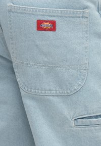 Υφασμα από ανοιχτό μπλε τζιν με τσέπη με πατς. Ετικέτα Dickies σε κόκκινο ραμμένη πάνω στην τσέπη. Απλός, ανθεκτικός σχεδιασμός με ορατές λεπτομέρειες ραφής.