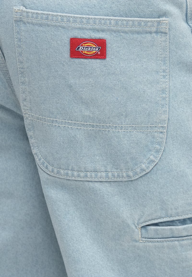 Hellblauer Denimstoff mit einer aufgesetzten Tasche. Rotes Dickies-Etikett auf der Tasche aufgenäht. Einfaches, strapazierfähiges Design mit sichtbaren Steppnähten.