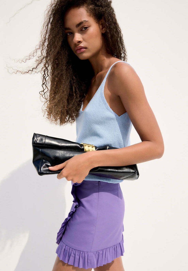 Femme aux cheveux bouclés portant un débardeur bleu clair et une jupe violette à volants, tenant une pochette noire avec un accent doré.