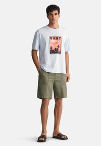 Camiseta de algodón azul claro con estampado gráfico y logotipo; pantalones cortos verdes de corte relajado con dos bolsillos frontales; sandalias de dedo negras.