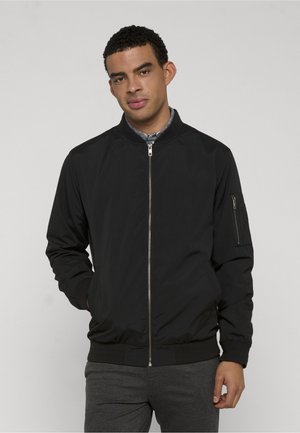 Młody mężczyzna w czarnej bomberce z zamkiem, na szarej koszuli zapinanej na guziki i ciemnoszarych spodniach, stojący na tle jednolitego, jasnego tła.