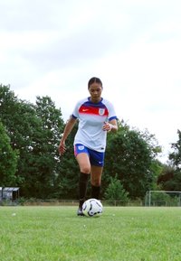 Jugador con uniforme blanco, rojo y azul de Nike con logo, calcetines negros hasta la rodilla, driblando un balón de fútbol blanco y negro en un campo de hierba.
