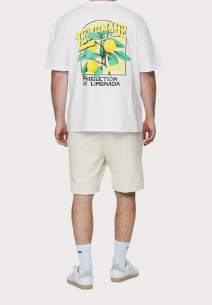 Wit T-shirt met korte mouwen, voorzien van een kleurrijke citroengrafiek en tekst op de achterkant, gecombineerd met lichte, gestructureerde shorts en witte sneakers.