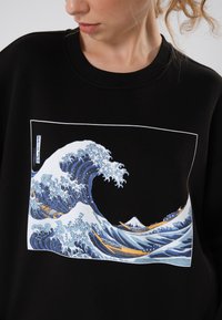Černá mikina s barevným motivem vlny v modrých a bílých odstínech, zobrazující lodě a horu uvnitř obdélníkového rámu.