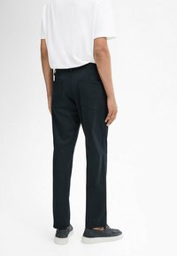 Black trousers with a straight fit, two back pockets, and a smooth texture; paired with a plain white shirt and gray shoes.