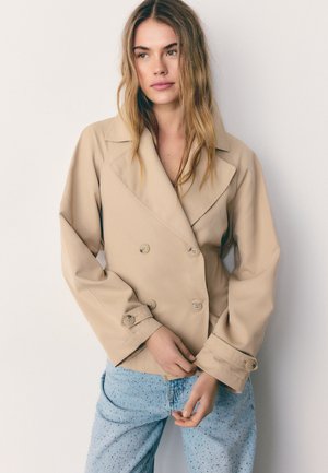 Jeune femme aux longs cheveux blonds portant une veste beige à double boutonnage et un jean bleu clair tacheté, debout devant un fond blanc.