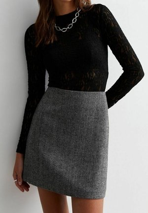Woman wearing a black lace long-sleeve top, silver chain necklace, and high-waisted gray herringbone skirt against a plain background.