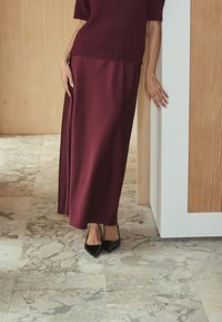 Burgundy dlouhá sukně z lehkého materiálu v kombinaci s tmavým pleteným topem. Na nohou jsou černé špičaté baleríny. Pozadí je tvořeno dřevěnými a kamennými texturami.