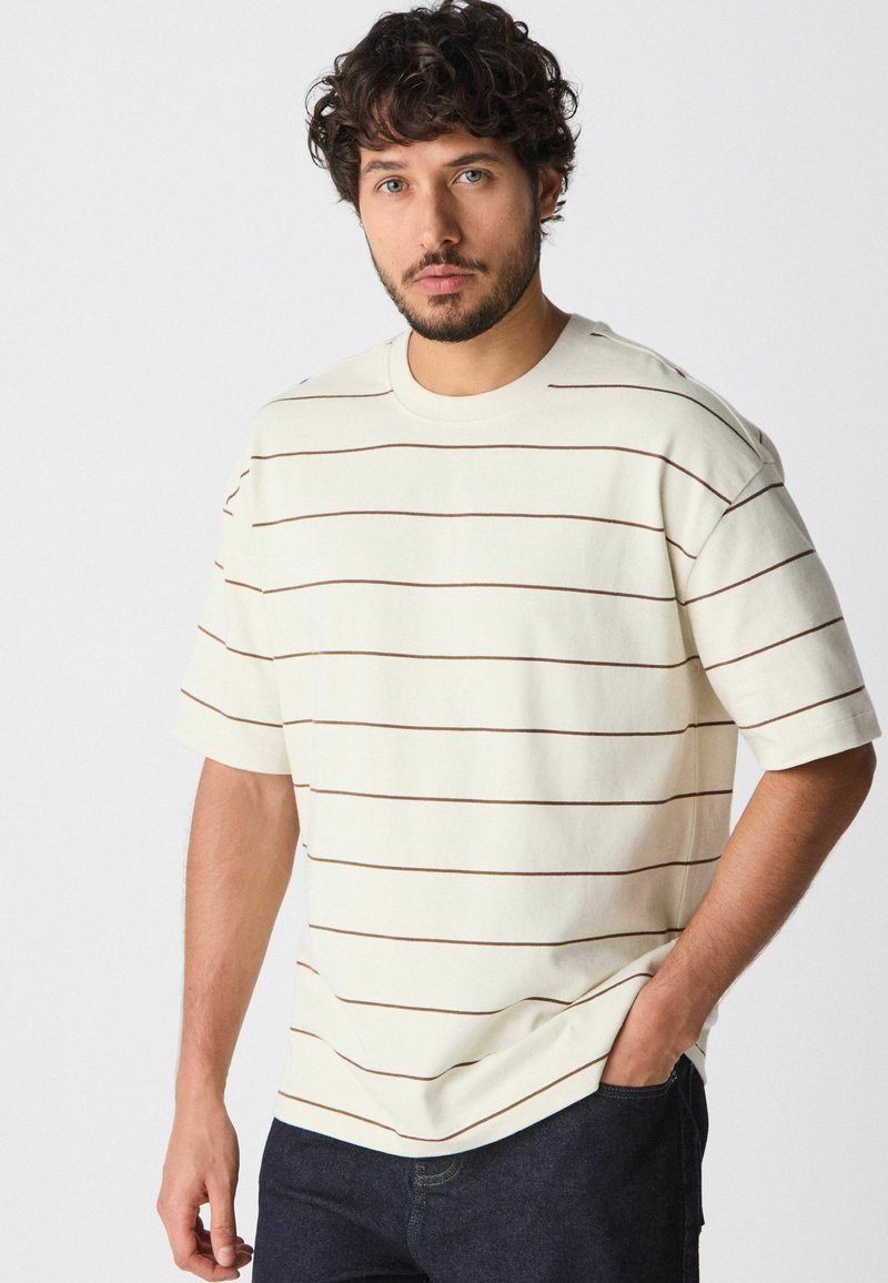 Crème T-shirt met korte mouwen en bruine horizontale strepen, gemaakt van zachte stof. Heeft een ronde hals en een relaxte pasvorm.
