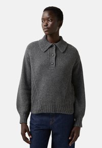 Vrouw in een grijze gebreide trui met een kraag en drie knopen, gecombineerd met donkerblauwe jeans, iets naar links gericht met een neutrale uitdrukking.