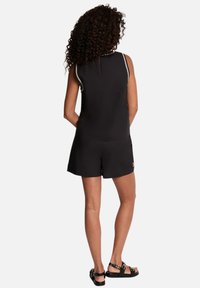 Mujer con cabello rizado que lleva una camiseta sin mangas negra y pantalones cortos a juego, de pie con las manos detrás de la espalda, con sandalias negras.