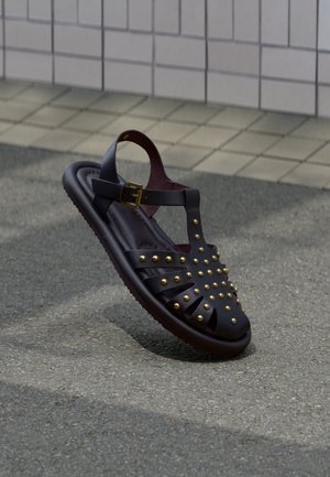 Sandalia de cuero negro con tachuelas doradas y una correa con hebilla, situada sobre una superficie gris texturizada cerca de un pavimento adoquinado.