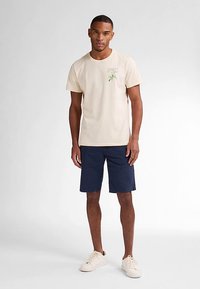 Beige puuvillainen t-paita, jossa kukkakirjailu, ja tummansiniset shortsit. Mies seisoo vaaleat tennarit jalassa yksivärisellä taustalla.