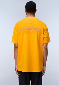 Žluté bavlněné tričko s kr�átkým rukávem, které na zadní straně zdobí velký grafický potisk s nápisem "NAPAPEURI geographic" v světle fialové barvě.